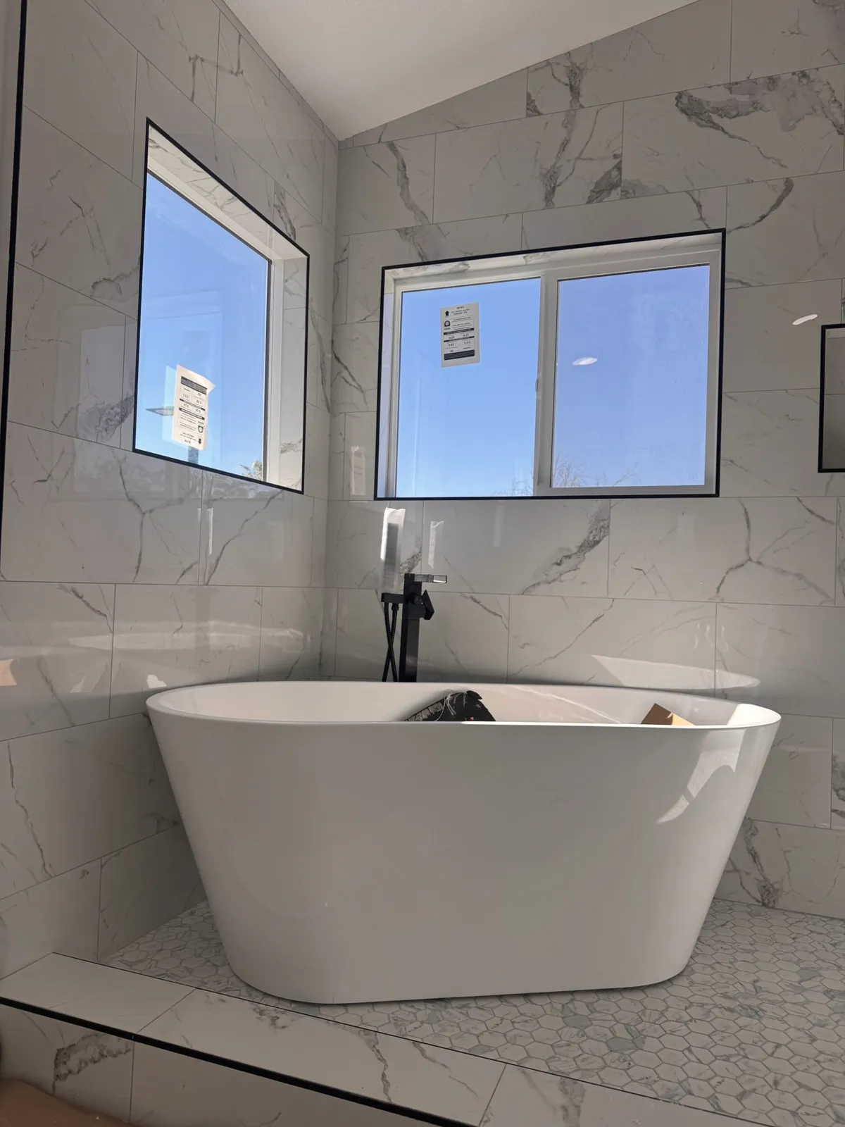 Freestanding white tub against marble-look tile walls under bright windows